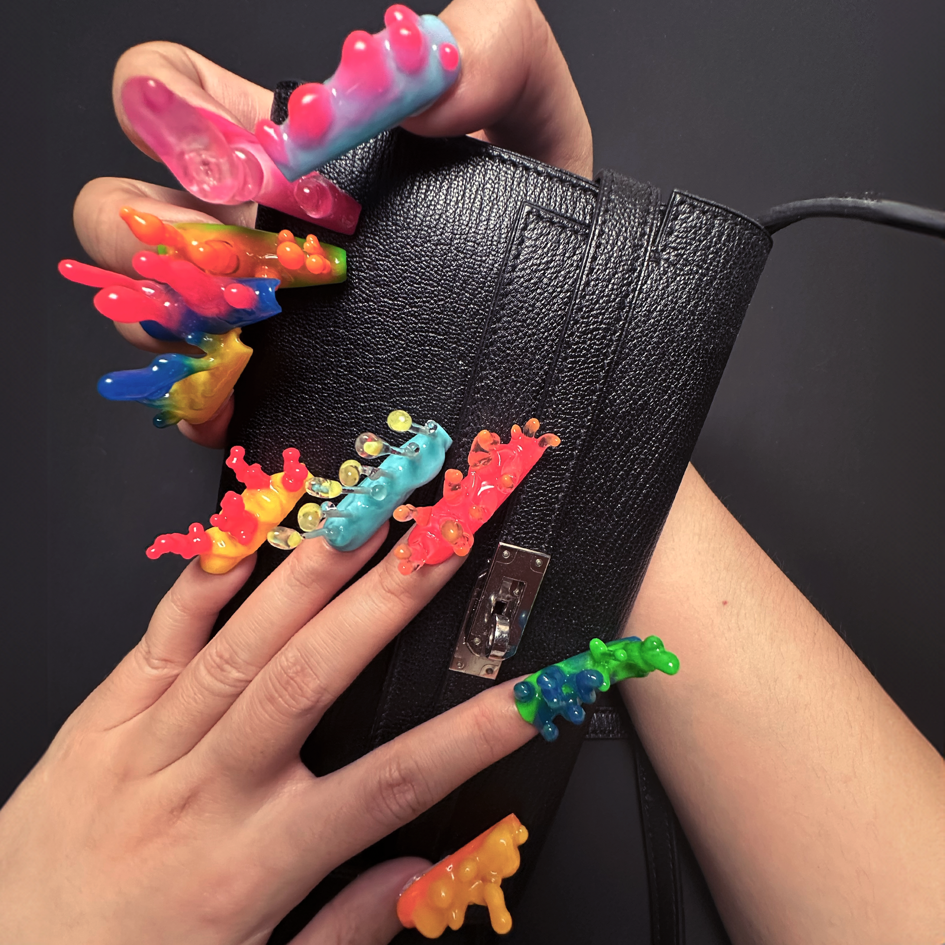 A close-up of hands displaying neon, handmade night glow 3D nails inspired by nudibranchs, featuring bright colors and bubble-like textures, holding a black Hermes handbag