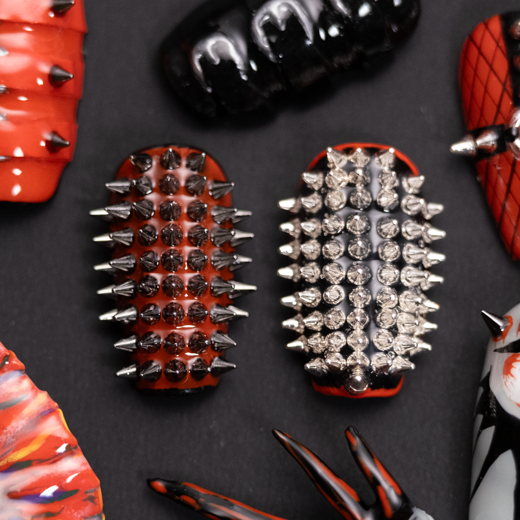 A close-up of two punk-inspired press-on nails, featuring bold red and black designs covered in metal spikes and studs for an edgy, rebellious look.