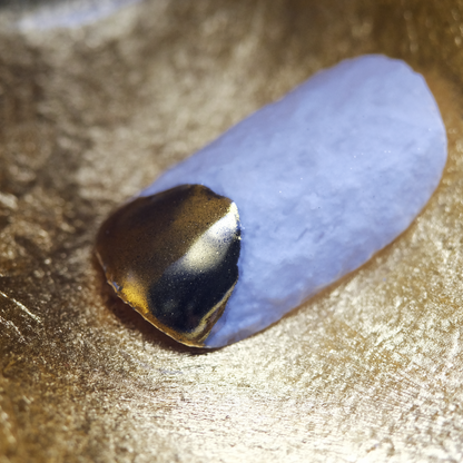 A close-up of a press-on nail with a textured white base and a gold tip, appearing as if dipped in gold, set against a metallic background.