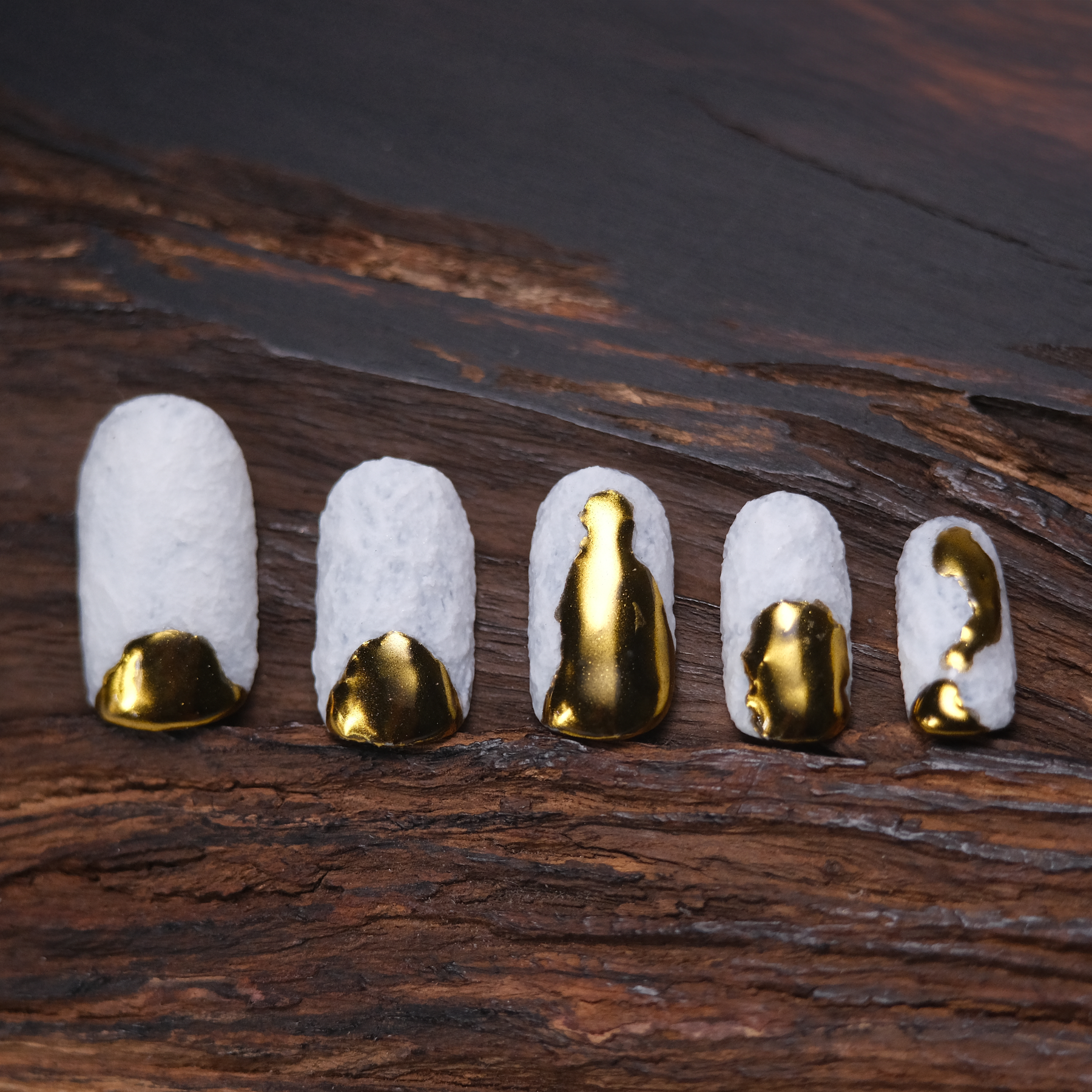 A set of press-on nails with a textured white base and irregular gold tips, appearing as if dipped in gold, set against a wooden background.