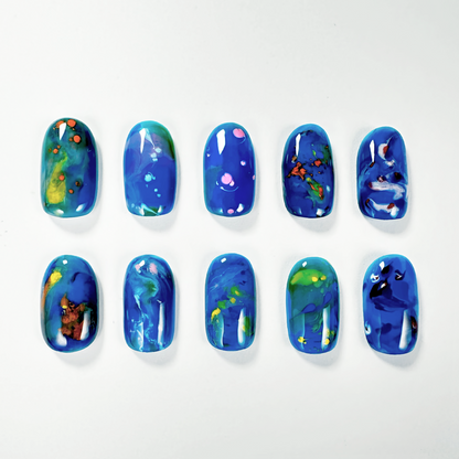 A set of nuance press-on nails featuring vibrant blue backgrounds with abstract designs in green, yellow, and pink, evoking a cosmic or oceanic theme.