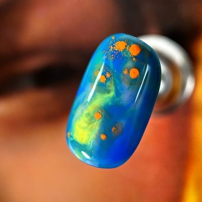 A close-up of a nuance press-on nail with a vibrant blue base, featuring dissolved swirls of green and yellow with orange specks, creating a cosmic effect.