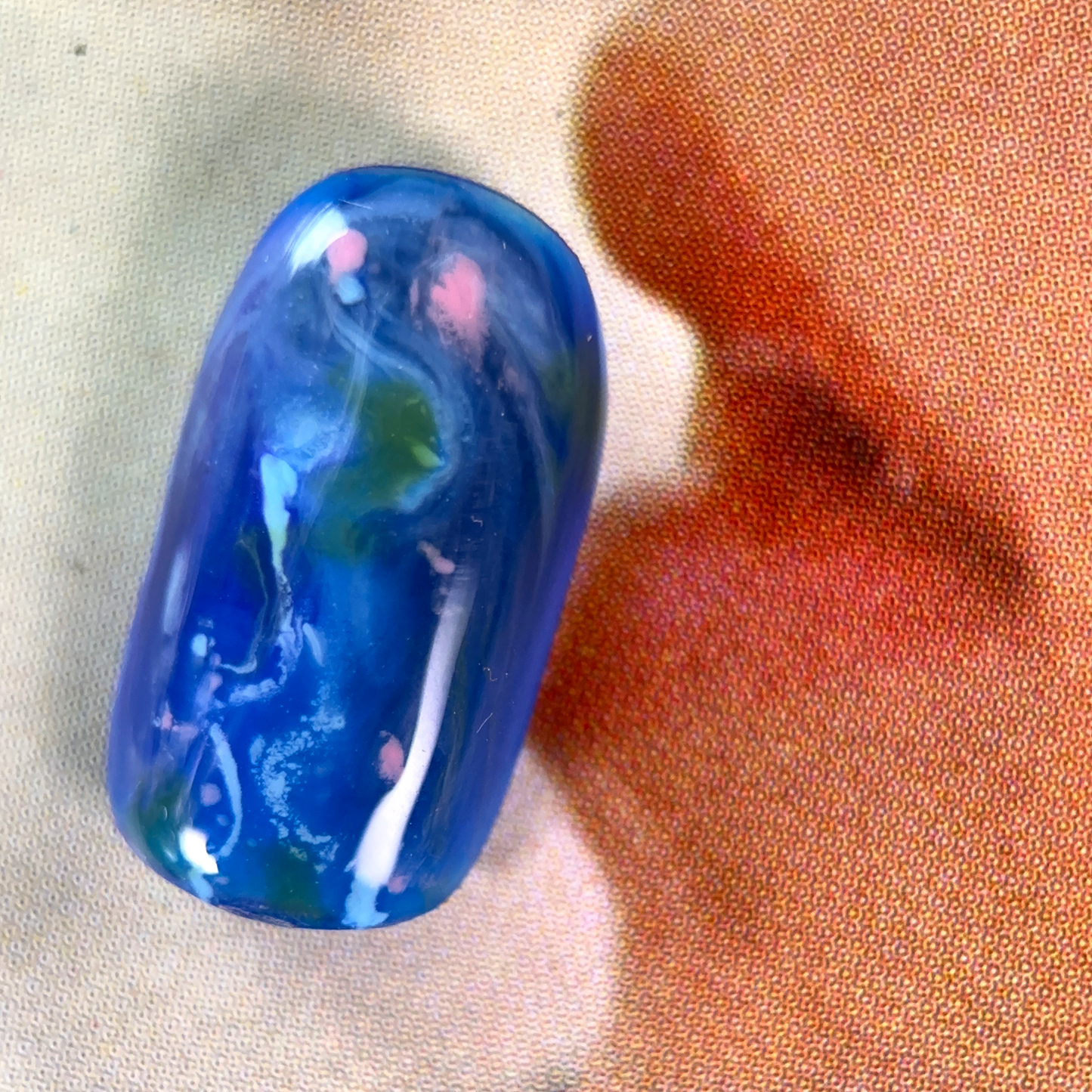 A close-up of a nuanced press-on nail with a deep blue base, featuring swirling patterns in green, pink, and white, creating an abstract, dreamy effect.