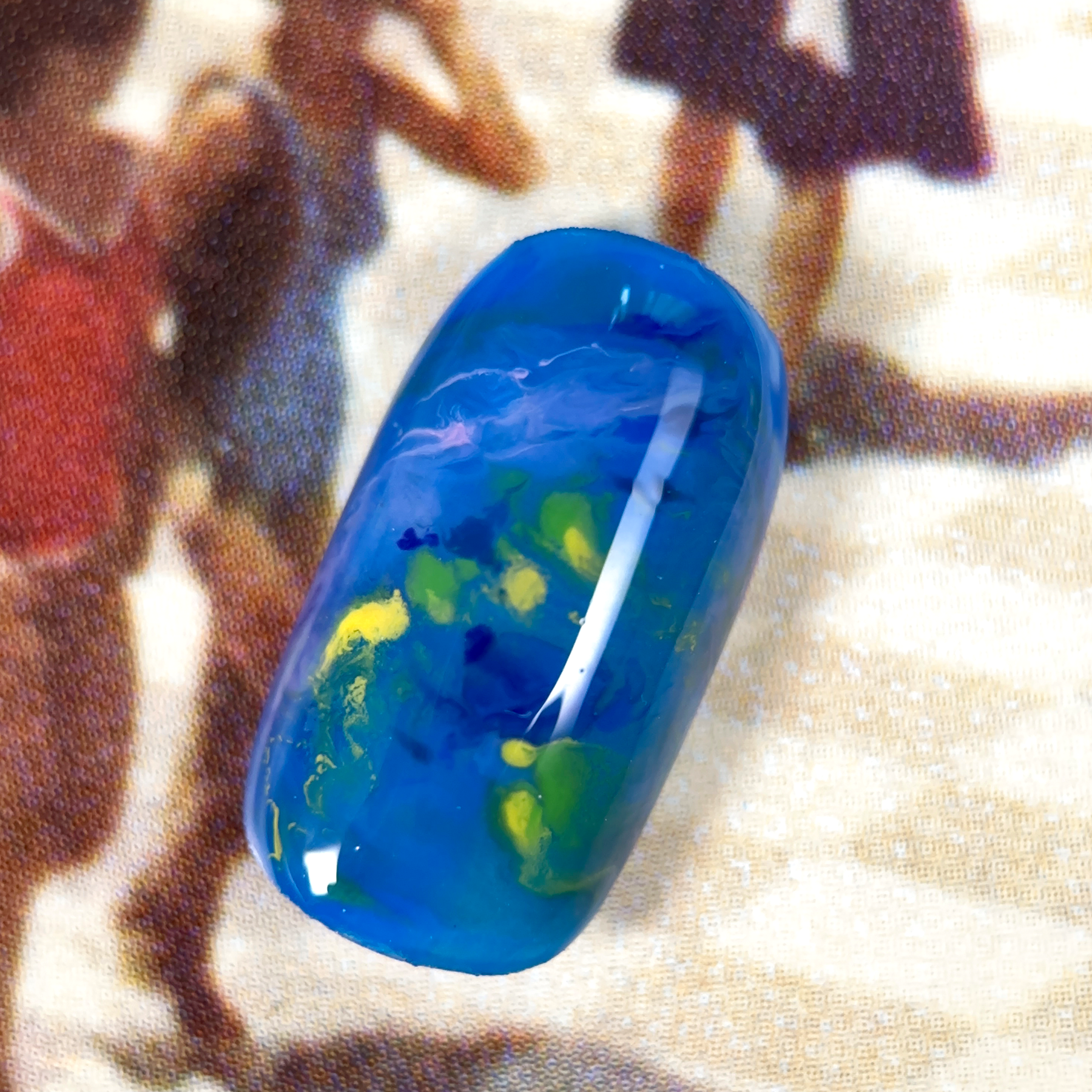 A close-up of a nuanced press-on nail with a deep blue base, featuring swirling green, yellow, and white tones, creating an abstract, oceanic effect.