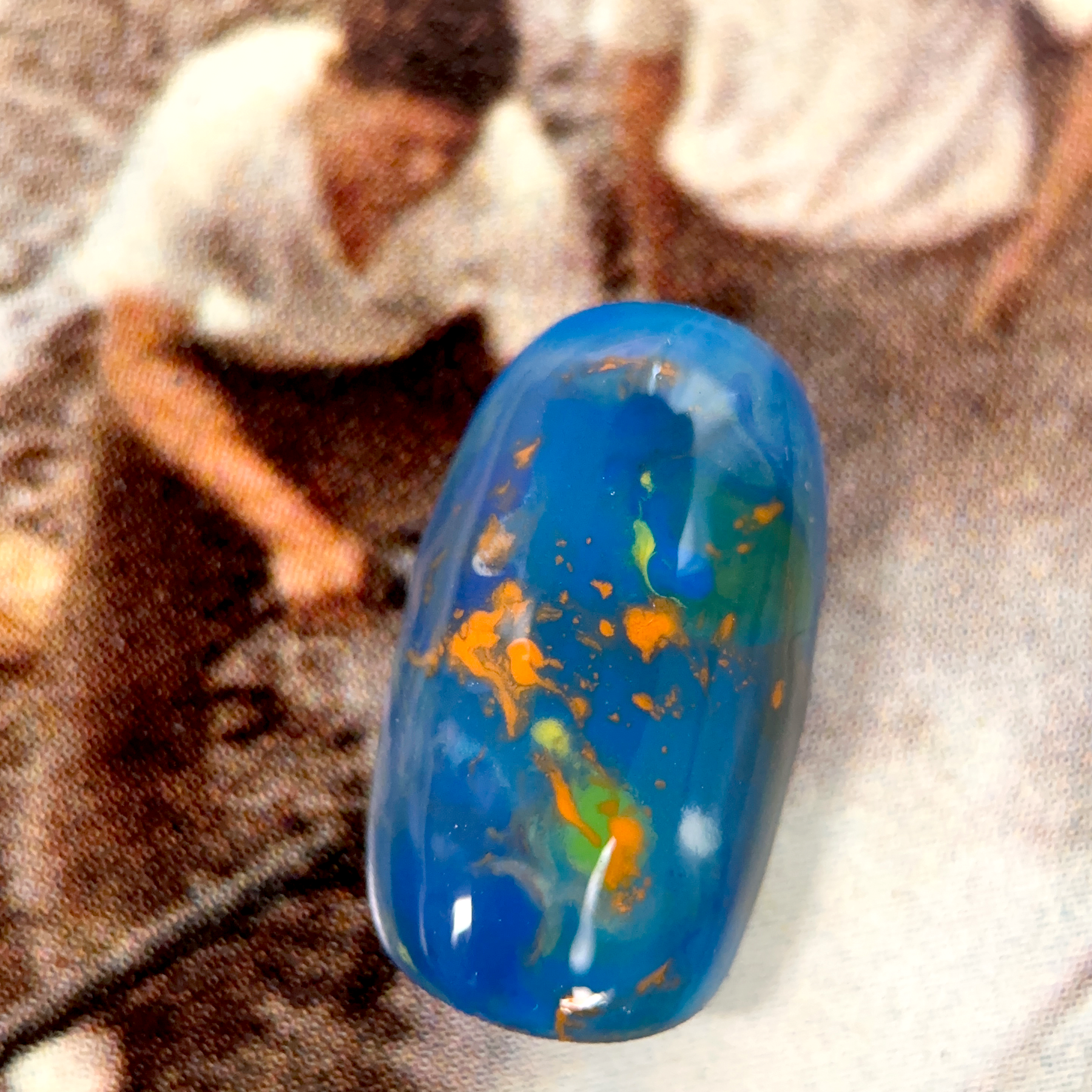 A close-up of a nuanced press-on nail with a deep blue base, featuring swirling orange and green tones, creating an abstract and vibrant effect.