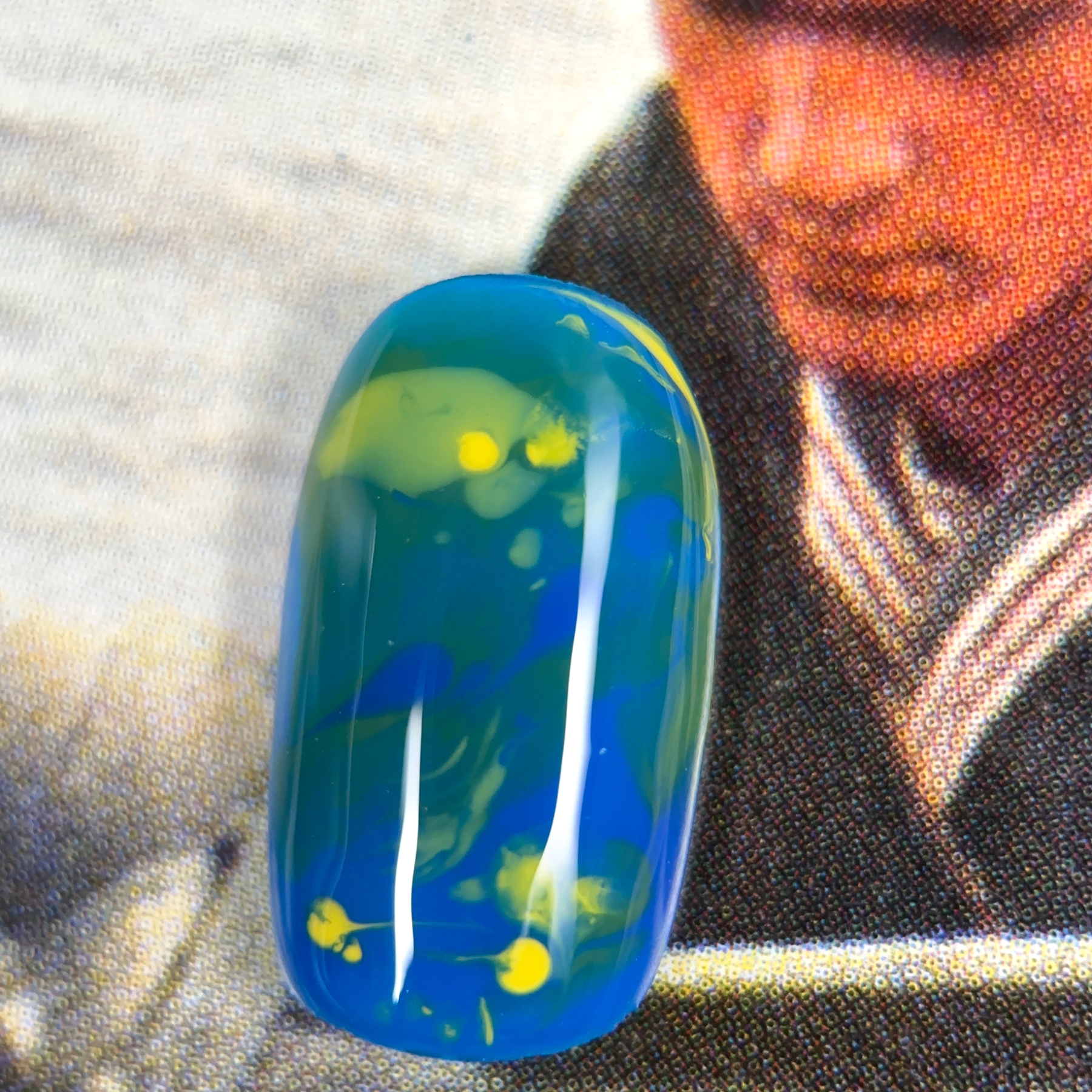 A close-up of a nuanced press-on nail with a deep blue base, featuring swirling yellow and green tones, creating an abstract, vibrant design.