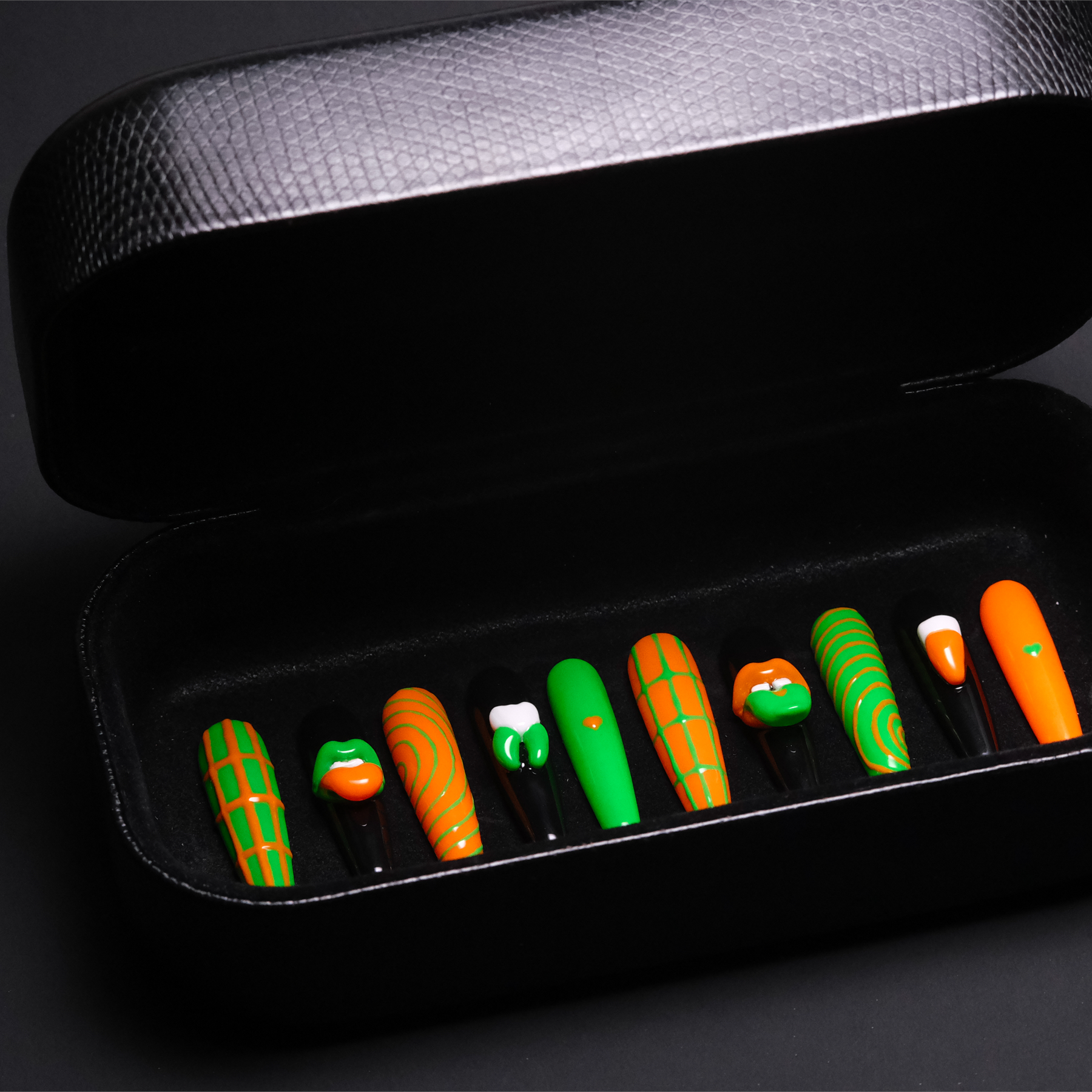 A set of vibrant press-on nails in a black case, showcasing a bold Gothic style with green and orange designs. The set features eye-catching 3D lip and tooth designs, enhancing the edgy, avant-garde look.